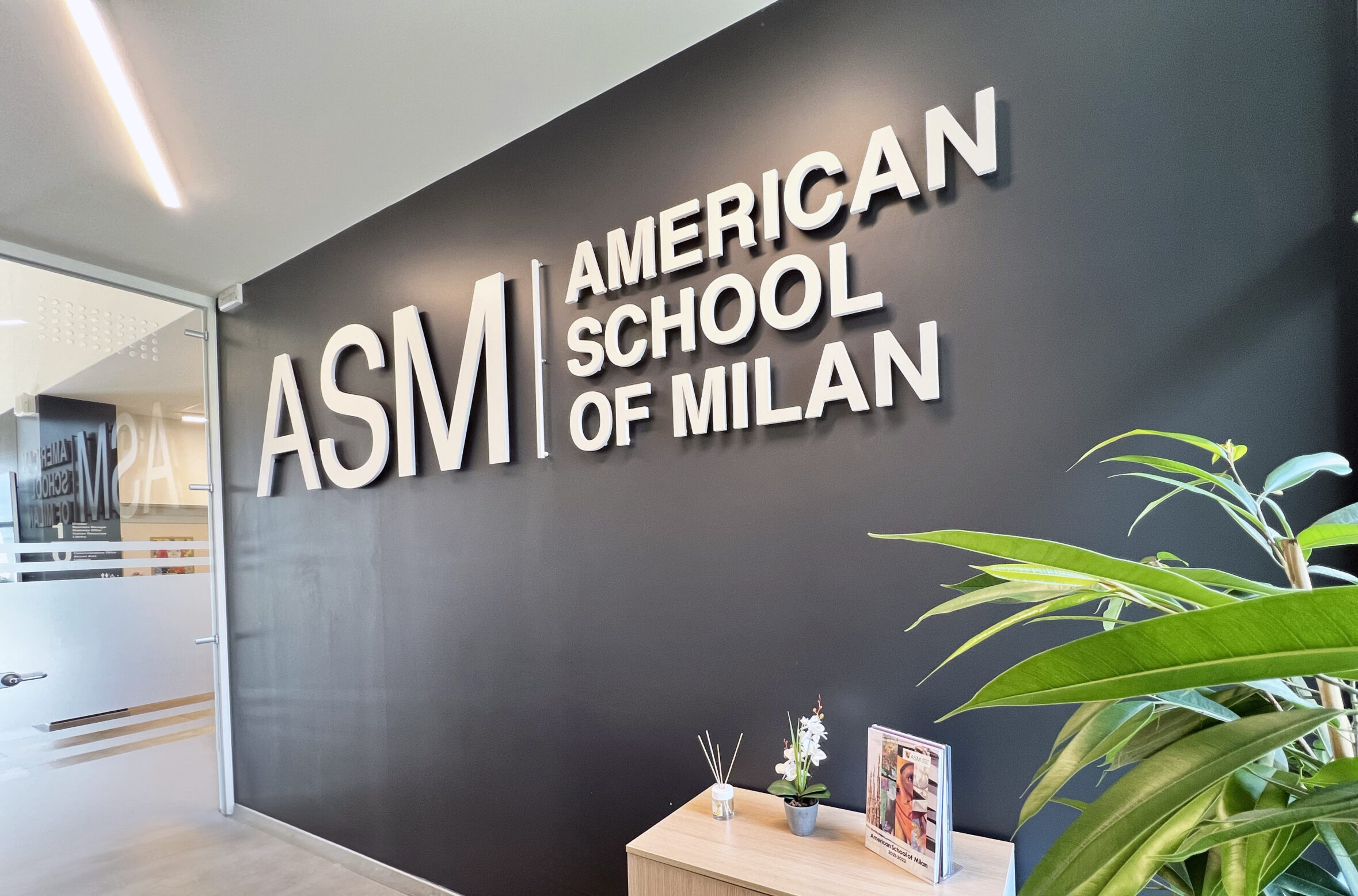 Interior lobby with a dark wall and large white 3D sign reading American School of Milan, visible beside a glass door and a reception desk with brochures and plants nearby.